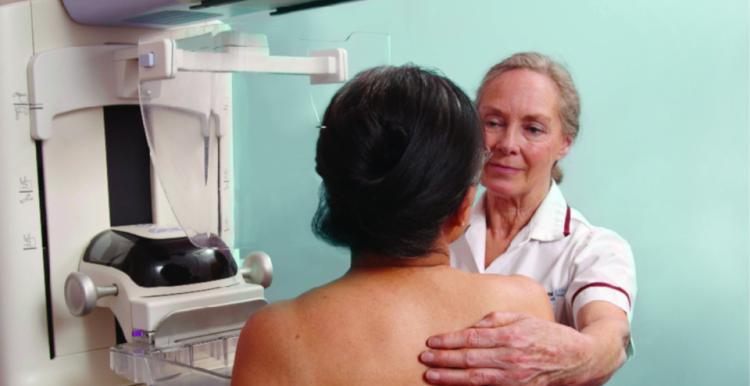 Woman standing next to mammography equipment, being helped into position by a female mammographer