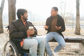 A man sitting in a wheelchair talking to another man on a bench