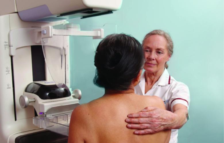 Woman standing next to mammography equipment, being helped into position by a female mammographer